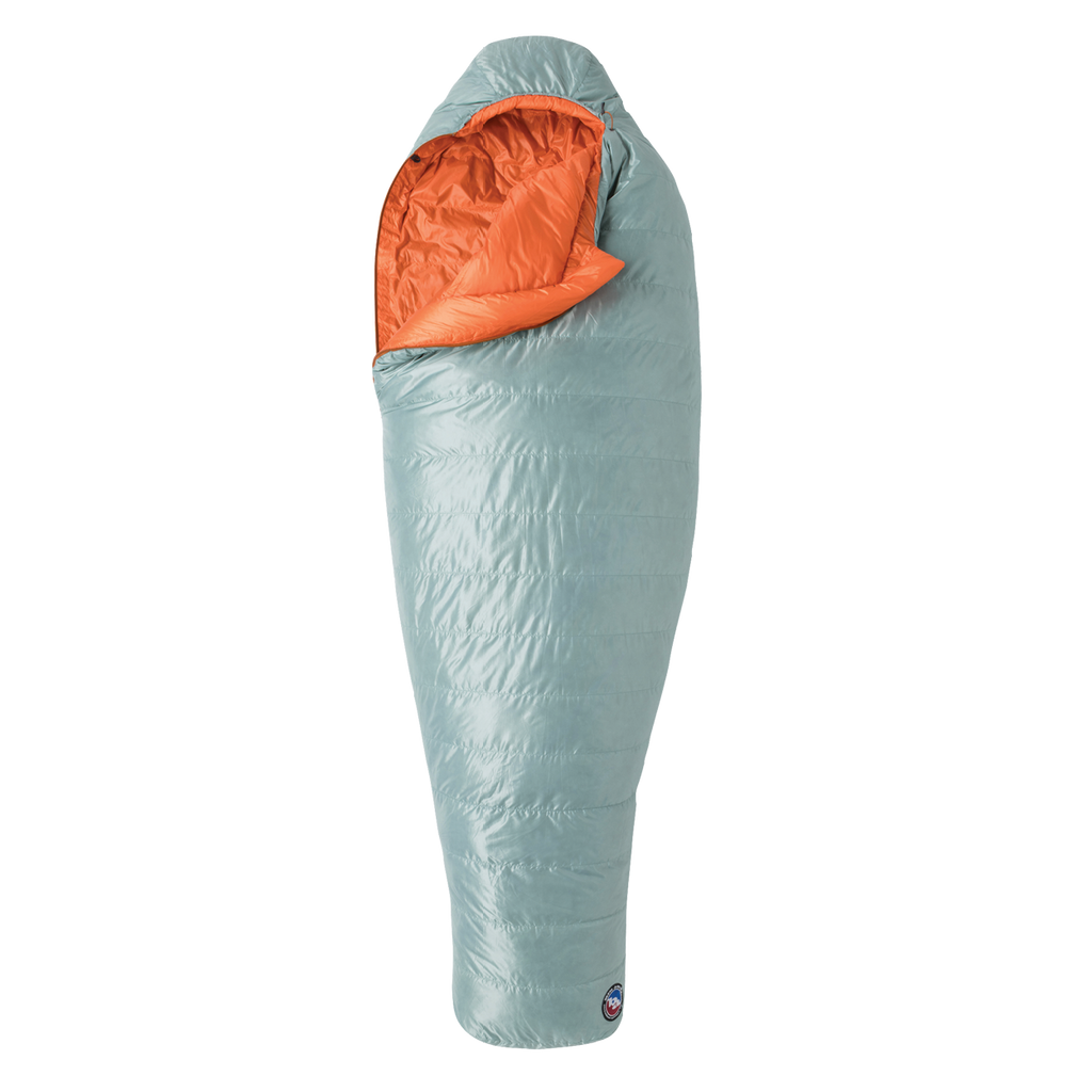 The Big Agnes Women's Greystone 30° sleeping bag in light blue, featuring DownTek insulation, stands upright against a white background. Its mummy-style hood is slightly open, showing the bright orange interior lining.